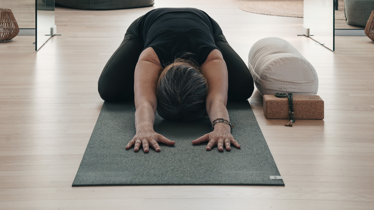 Kvinde i barnets stilling på en grøn yogamåtte med bolster, yogablok i kork og mala